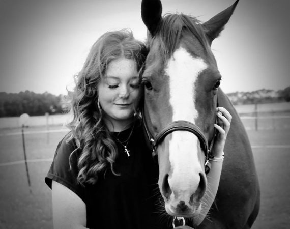 Young woman with long curly hair stands beside a horse, resting her head against its neck in a calm field.