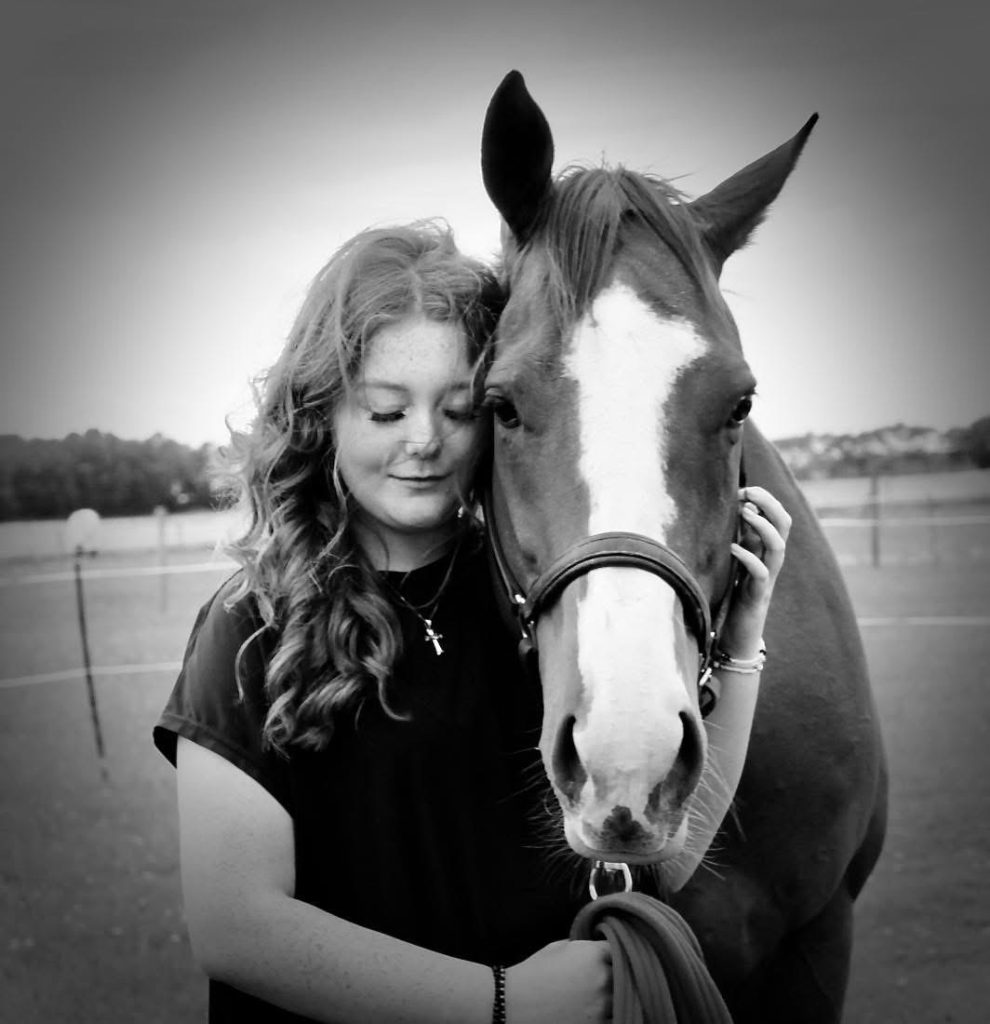 Young woman with long curly hair stands beside a horse, resting her head against its neck in a calm field.