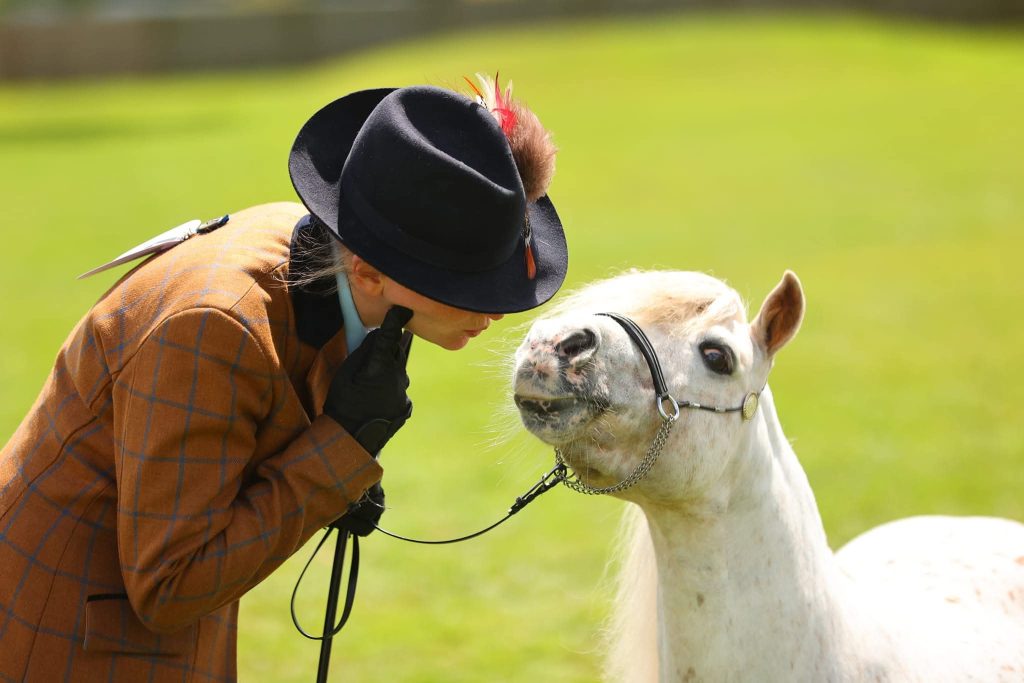 Person in a brown plaid jacket and black hat leaning toward a white horse in a green field outdoors.